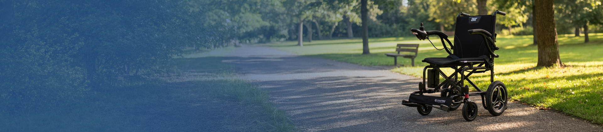 A black electric wheelchair sits empty on a paved path in a sunny park, surrounded by green grass and trees, with a wooden bench visible in the background.