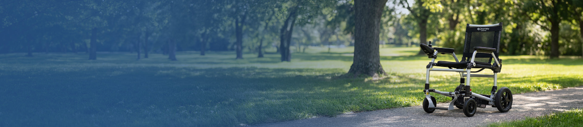 A lightweight Certified Pre-Owned electric wheelchair sits empty on a paved path in a sunny park, surrounded by green grass and trees.