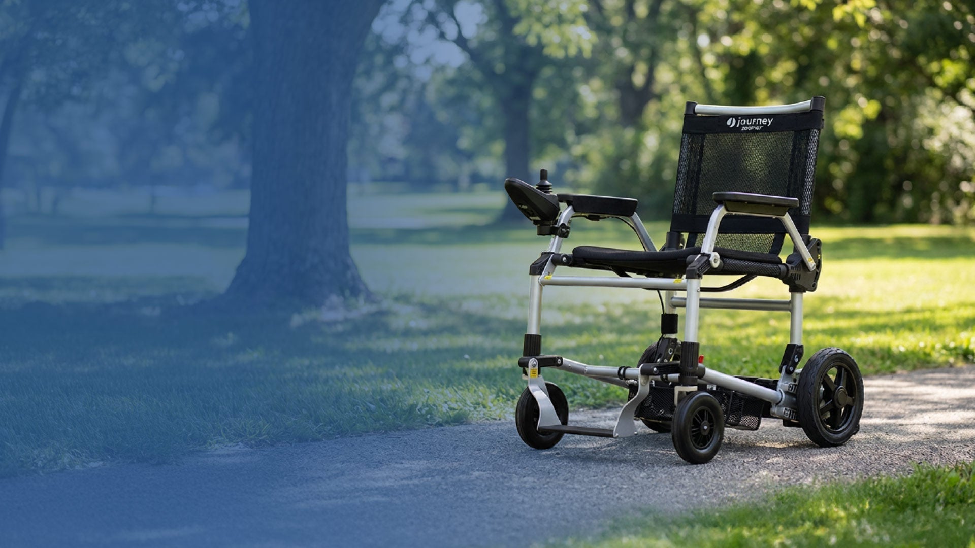 A lightweight, black electric wheelchair sits empty on a paved path in a sunlit park, with trees and grass in the background.