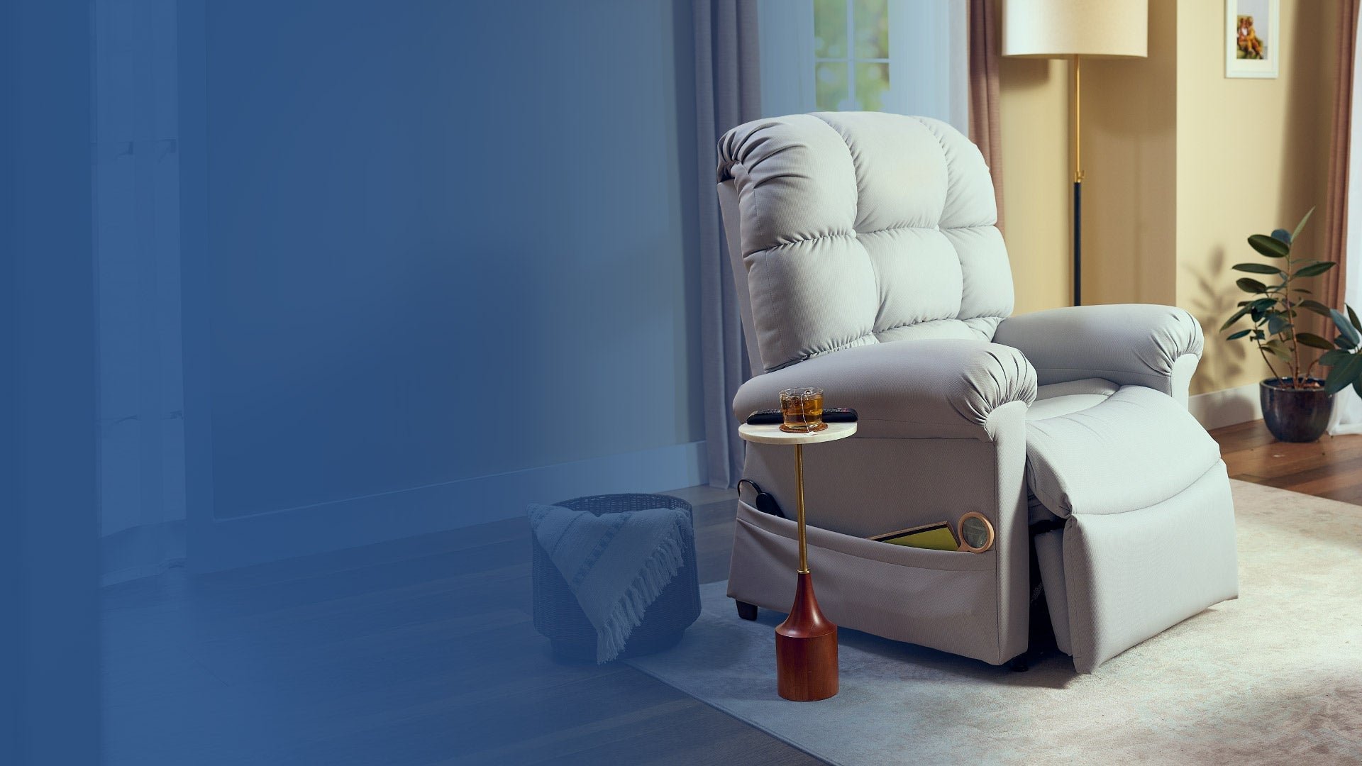 A plush gray recliner sits next to a small side table with a drink and book on it, in a cozy living room with a blanket basket, plant, floor lamp, and light streaming through the window.
