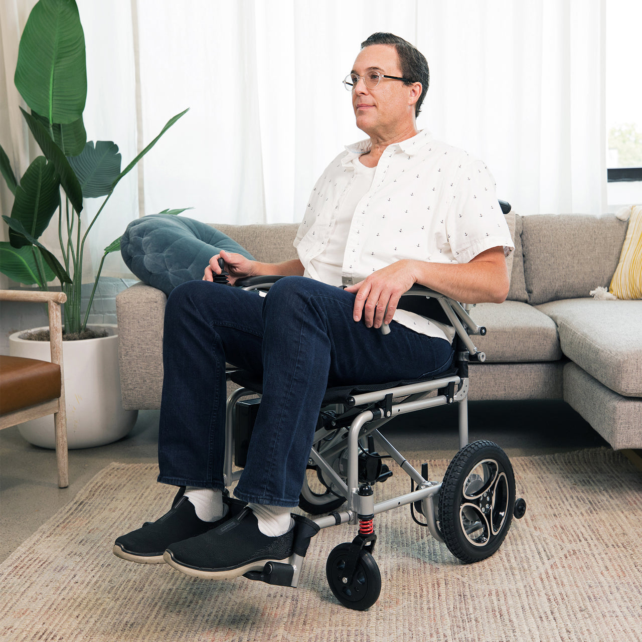 A man in glasses, a white shirt, and jeans sits on the Journey Air Lightweight Folding Power Chair with dual brushless motors in a living room featuring a beige sofa, potted plant, and light-colored rug.