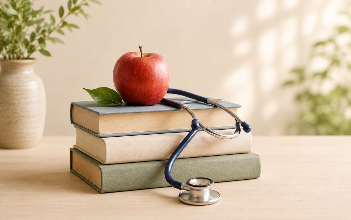 A red apple with a green leaf rests on a stack of three books, with a blue stethoscope draped over the books on a light wooden table. The background is softly blurred with green plants and warm sunlight.