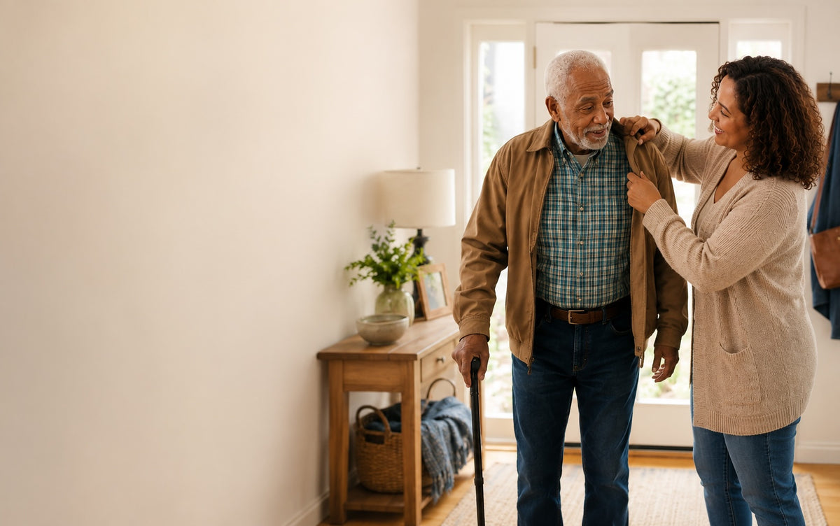 An elderly man with a cane uses a walker while a woman adjusts his jacket in a brightly lit home interior.