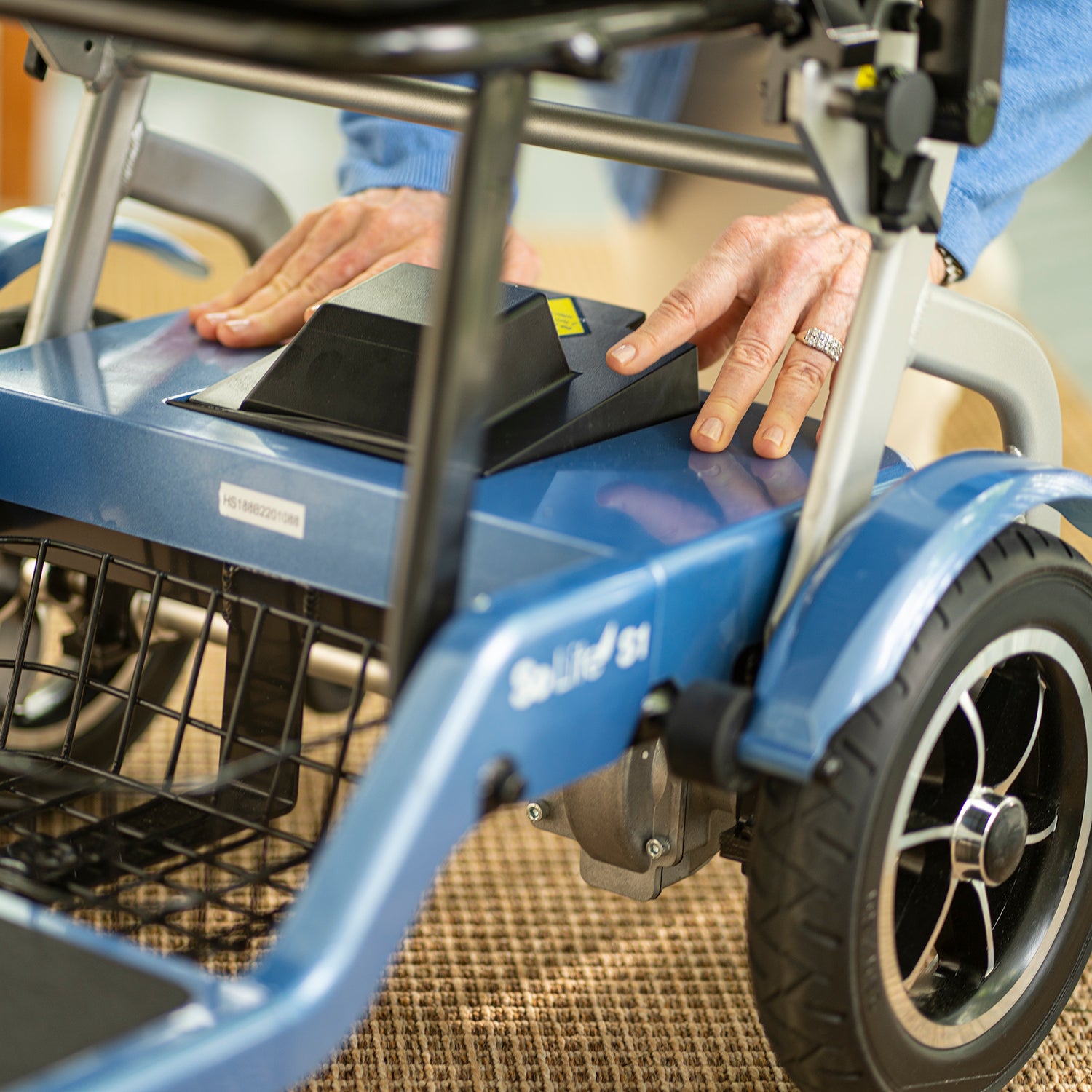Close-up of hands adjusting a black part on the Journey So Lite Folding Power Scooter, highlighting its blue frame, rear wheel, and storage basket. The user wears a silver ring and a blue sleeve.