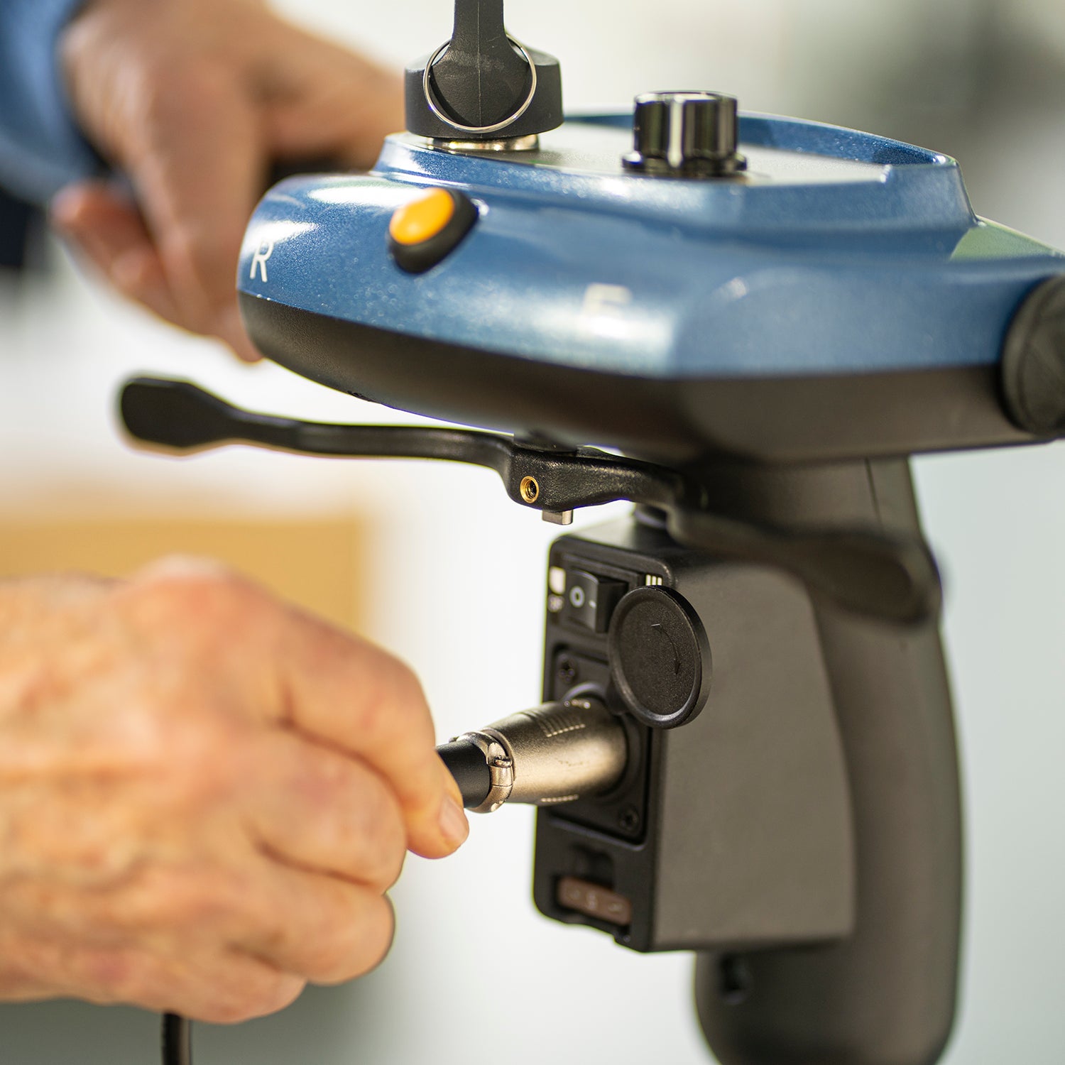 Close-up of a hand connecting a cable to the blue and black control panel—featuring buttons, levers, and joystick—of the Journey So Lite Folding Power Scooter, a lightweight model with electronic stability control.