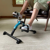 A person uses the Journey Exercise Peddler for low-impact workouts while seated on a chair, with the device placed on a light-colored carpet in a room featuring wooden trim and a window in the background.