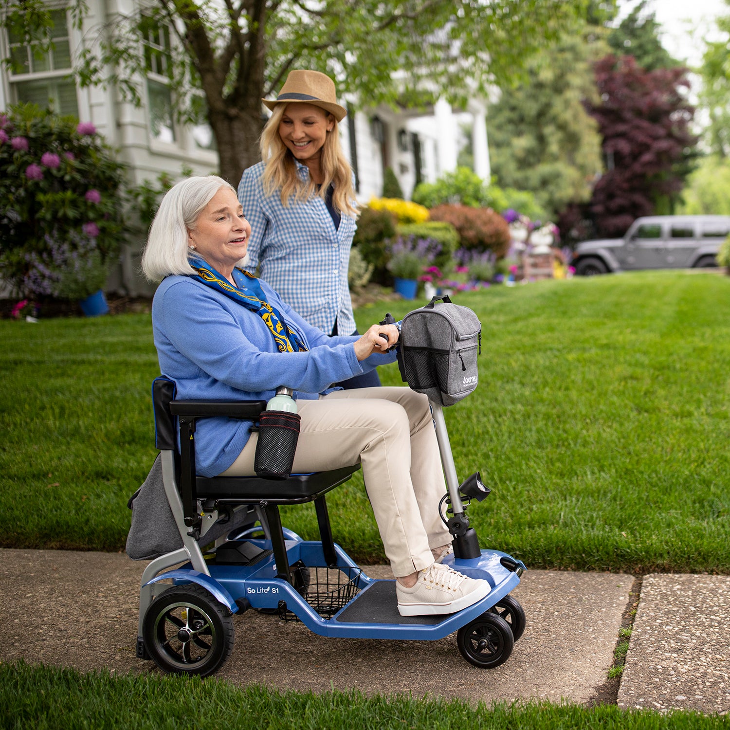 An older woman in a blue sweater smiles while sitting on the Journey So Lite Folding Power Scooter beside a woman in a hat. They are outdoors on a sidewalk in front of a house with green lawn and blooming flowers.