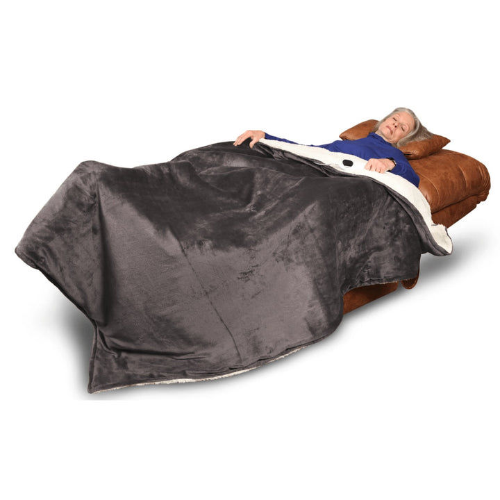 An older woman relaxes on a brown recliner, covered with a large gray Journey Companion Heating Blanket, looking comfortable and at ease.