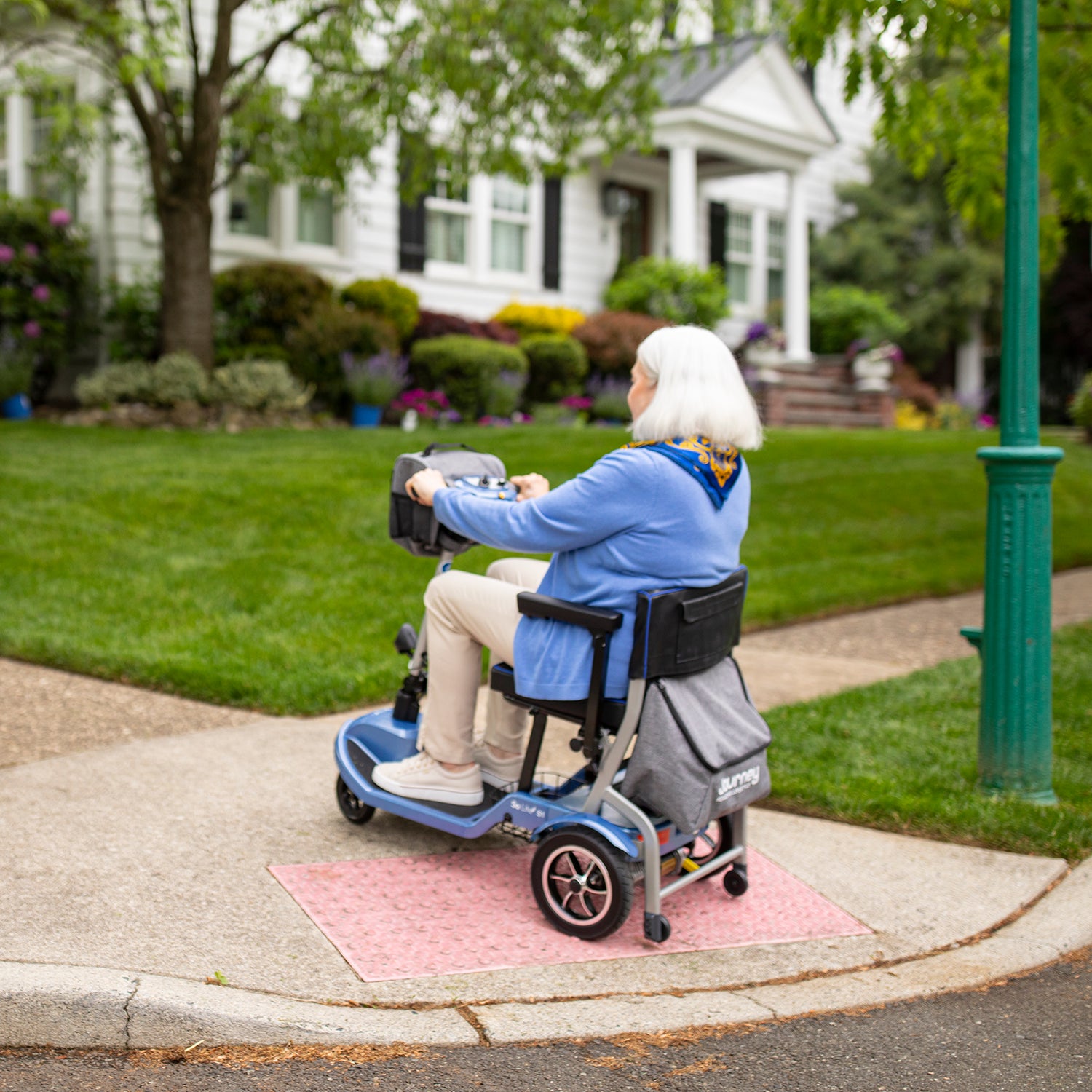 An older woman with white hair rides a Journey So Lite Folding Power Scooter along a sidewalk near a green lawn and white house, passing by a green streetlamp.