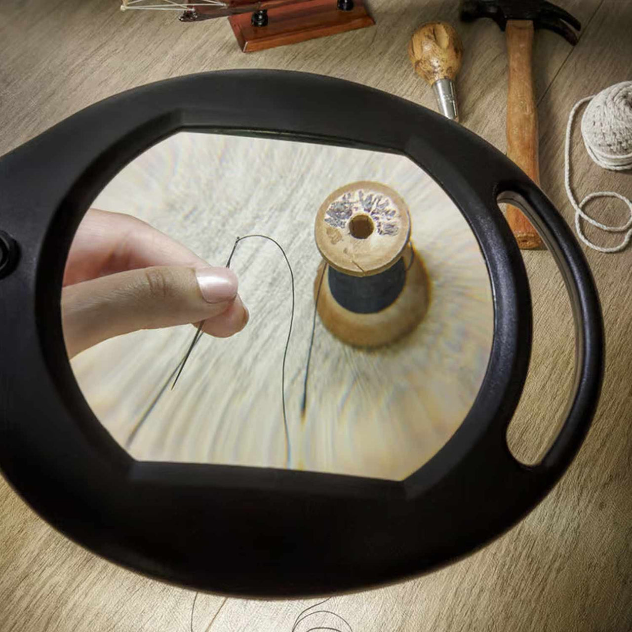 A hand threading a needle is seen through the 5X Glass Floor Stand Magnifier With Light, with a spool of thread, hammer, and wooden-handled tool placed on the wooden table in the background.