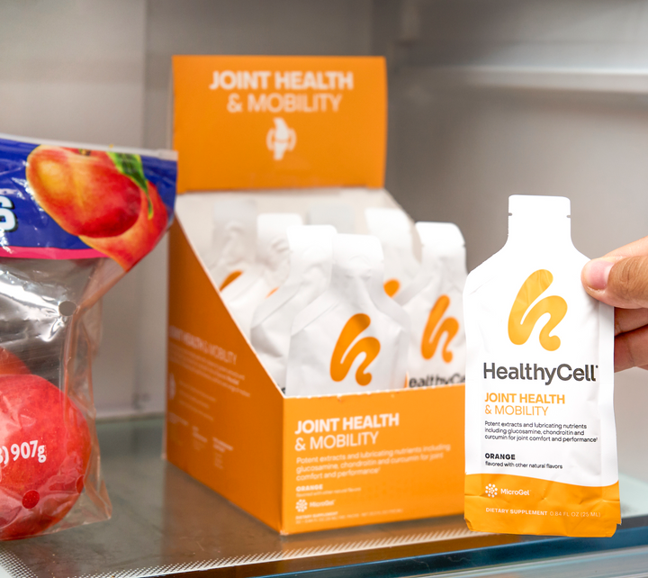 A hand holds a Joint Health & Mobility Supplement pouch in front of an open refrigerator box filled with similar pouches, next to a bag of red apples.