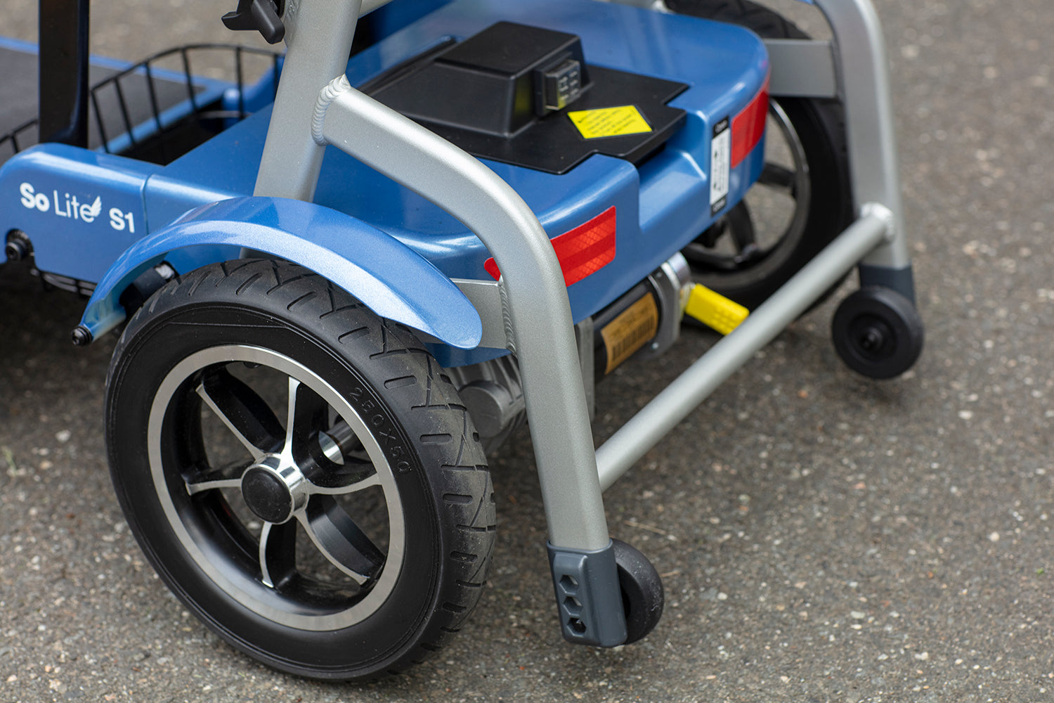 Close-up of the rear wheel and frame of the blue Journey So Lite Folding Power Scooter, featuring black tires, red reflectors, stabilizer wheels, and electronic stability control on a paved surface.