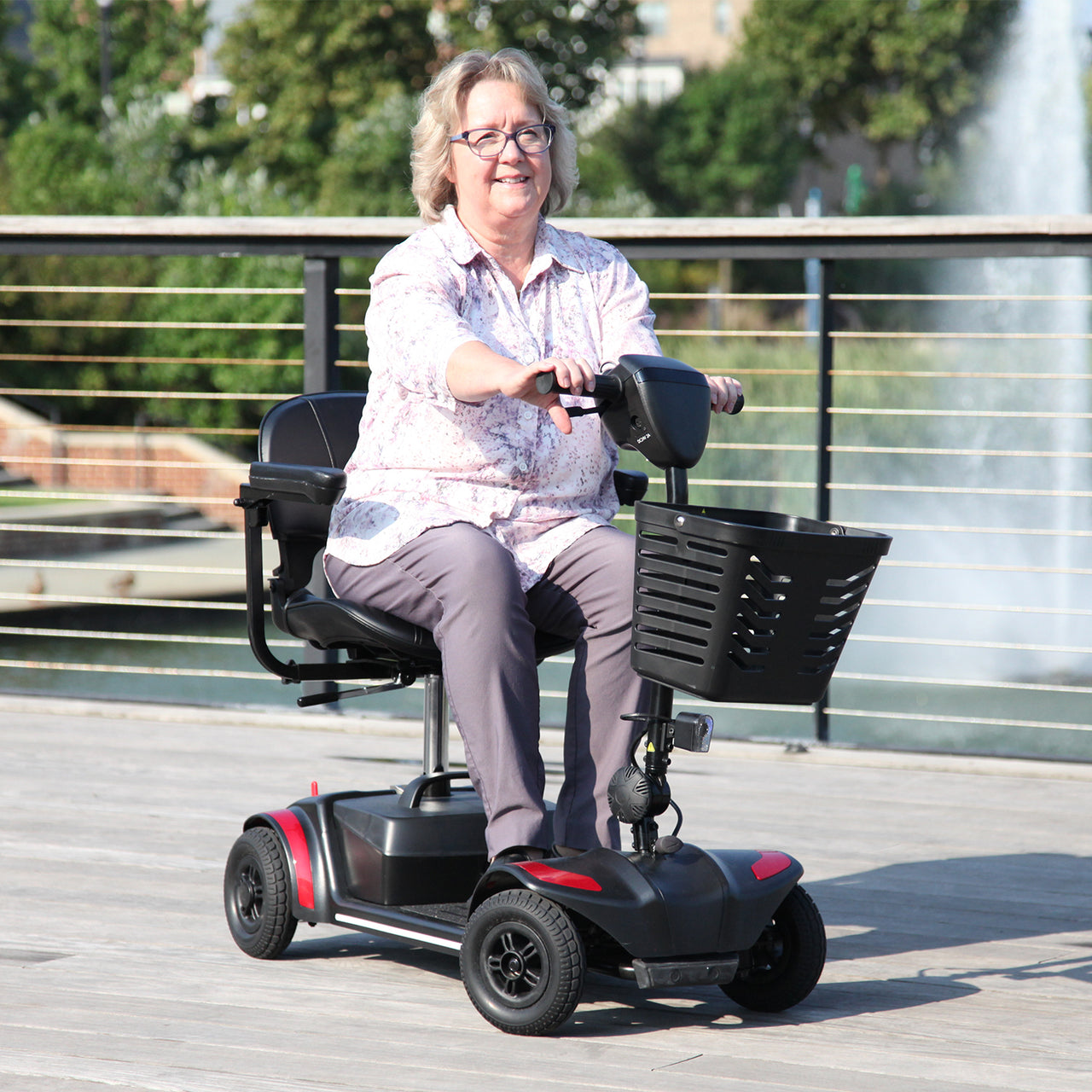 A smiling older woman with glasses rides the Journey Adventure Scooter on a wooden boardwalk outdoors, enjoying its effortless portability amid green trees and water in the background.