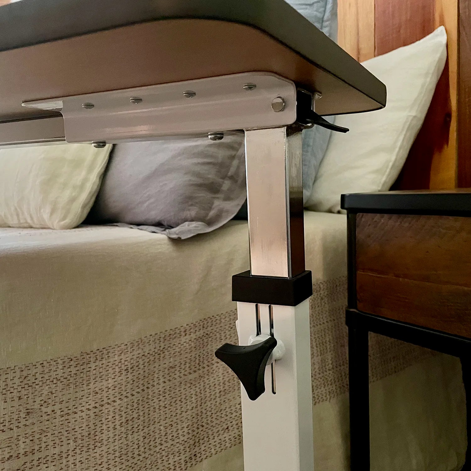 Close-up of the Journey™ Overbed Table – Adjustable & Portable Design by a bed with pillows and a nightstand, highlighting its height adjustment mechanism and clamp—perfect as a rolling overbed table for home recovery.