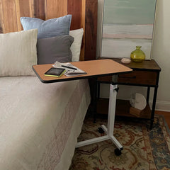 The Journey Overbed Table – Adjustable & Portable Design sits next to a bed adorned with pillows and a nightstand with decor in a cozy bedroom. Topped with a Kindle, reading glasses, and paper, this hospital-style rolling bedside table features an easy-clean tabletop, perfectly blending practicality with comfort.