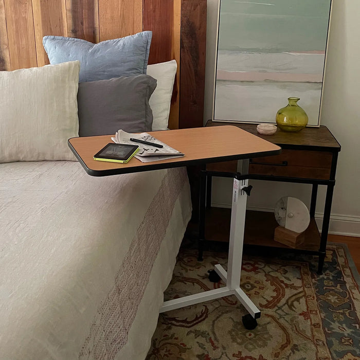 The Journey Adjustable Height Overbed Table with swivel casters is placed over a bed with pillows, holding a Kindle, eyeglasses, and papers. Next to the bed is a nightstand with decorative items and a framed painting.