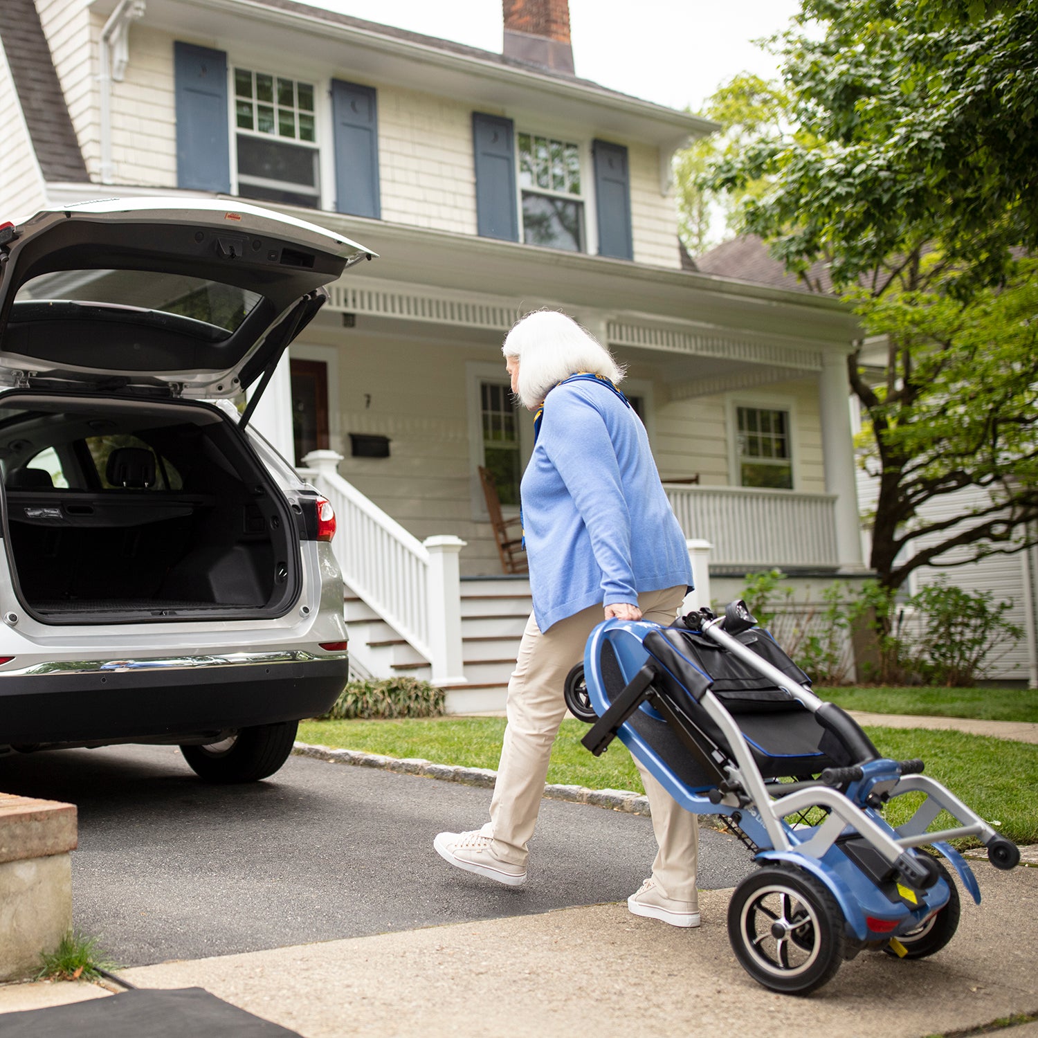 An older woman with gray hair, in a blue sweater and beige pants, walks toward an open car trunk while carrying a Journey So Lite Folding Power Scooter in the driveway of a house.