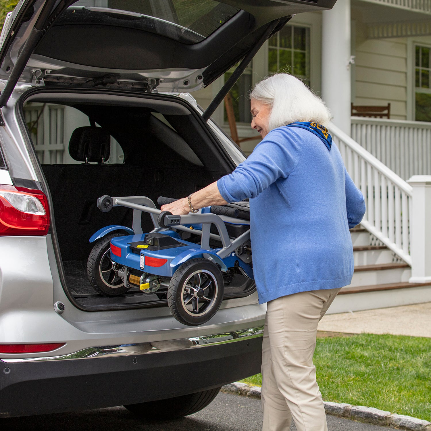 An older woman with gray hair loads a folded Journey So Lite Folding Power Scooter into the trunk of an SUV parked outside a house with a porch and white railing.