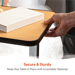 A hand presses down on the corner of a wooden tabletop holding a closed book. Text below reads, Secure & Sturdy. Keep your Journey Overbed Table – Adjustable & Portable Design securely in place with its lockable tabletop.