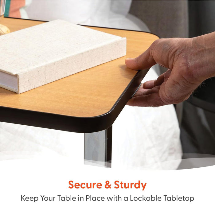 A hand presses down on the corner of a wooden tabletop holding a closed book. Text below reads, Secure & Sturdy. Keep your Journey Overbed Table – Adjustable & Portable Design securely in place with its lockable tabletop.