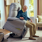 An older man smiles as he uses the remote of the Perfect Sleep Chair & Lift Chair, which is tilted forward to assist him in standing up. He sits in a bright, cozy living room with large windows and beige decor.