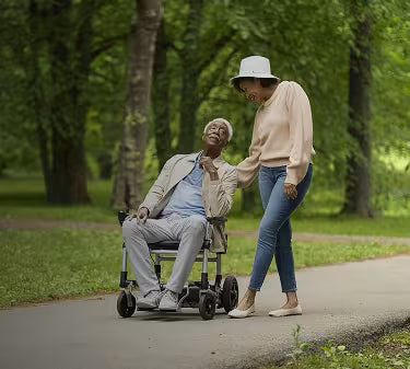 An elderly man in a light beige blazer and gray trousers sits in a motorized wheelchair on a paved path in a park, while an elderly woman wearing a beige sweater, blue jeans, and a white hat stands beside him.