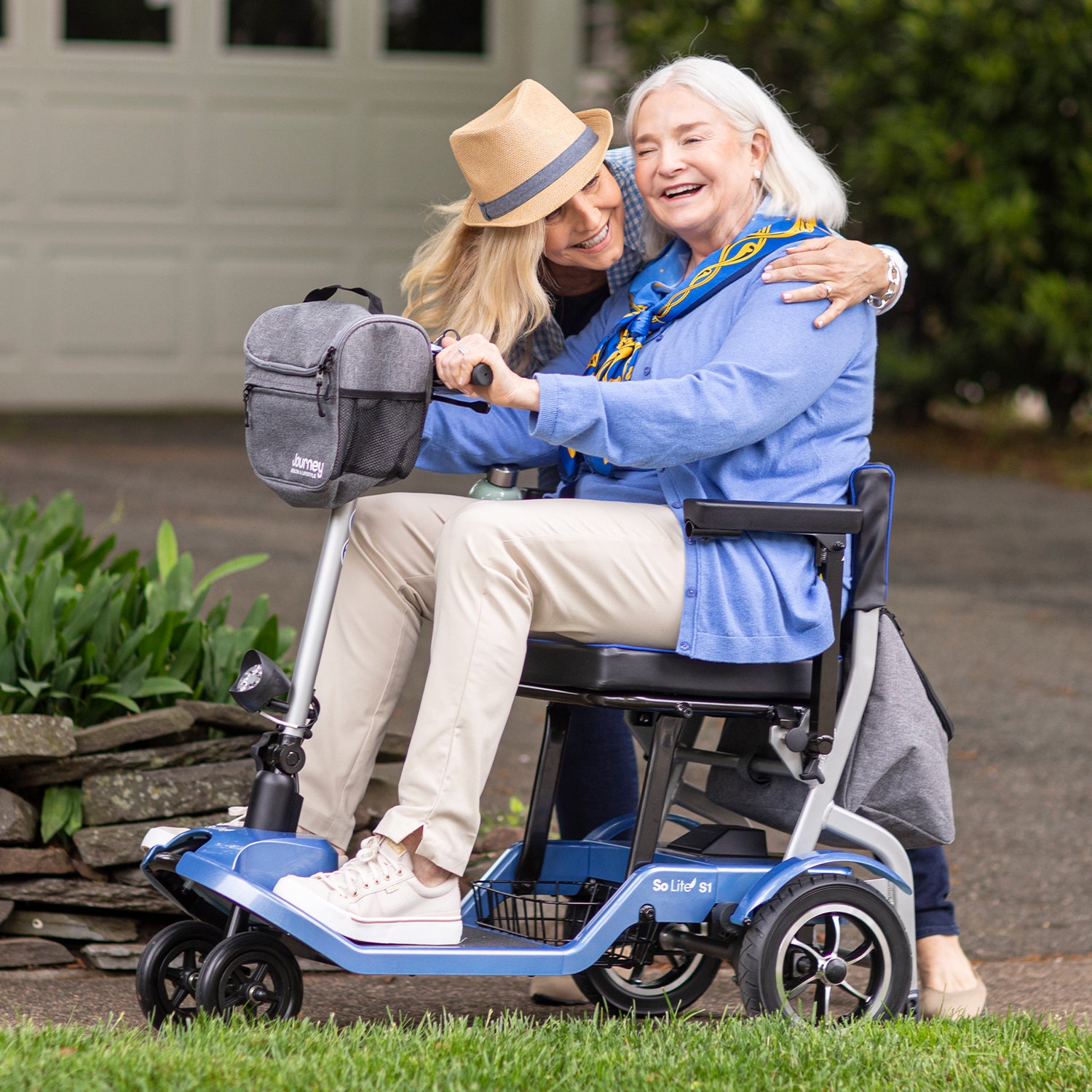 An older woman, smiling and joyful in blue, sits outdoors on a Journey So Lite Folding Power Scooter as she embraces a younger, hat-wearing companion; together they enjoy their time and the scooter’s lightweight convenience.