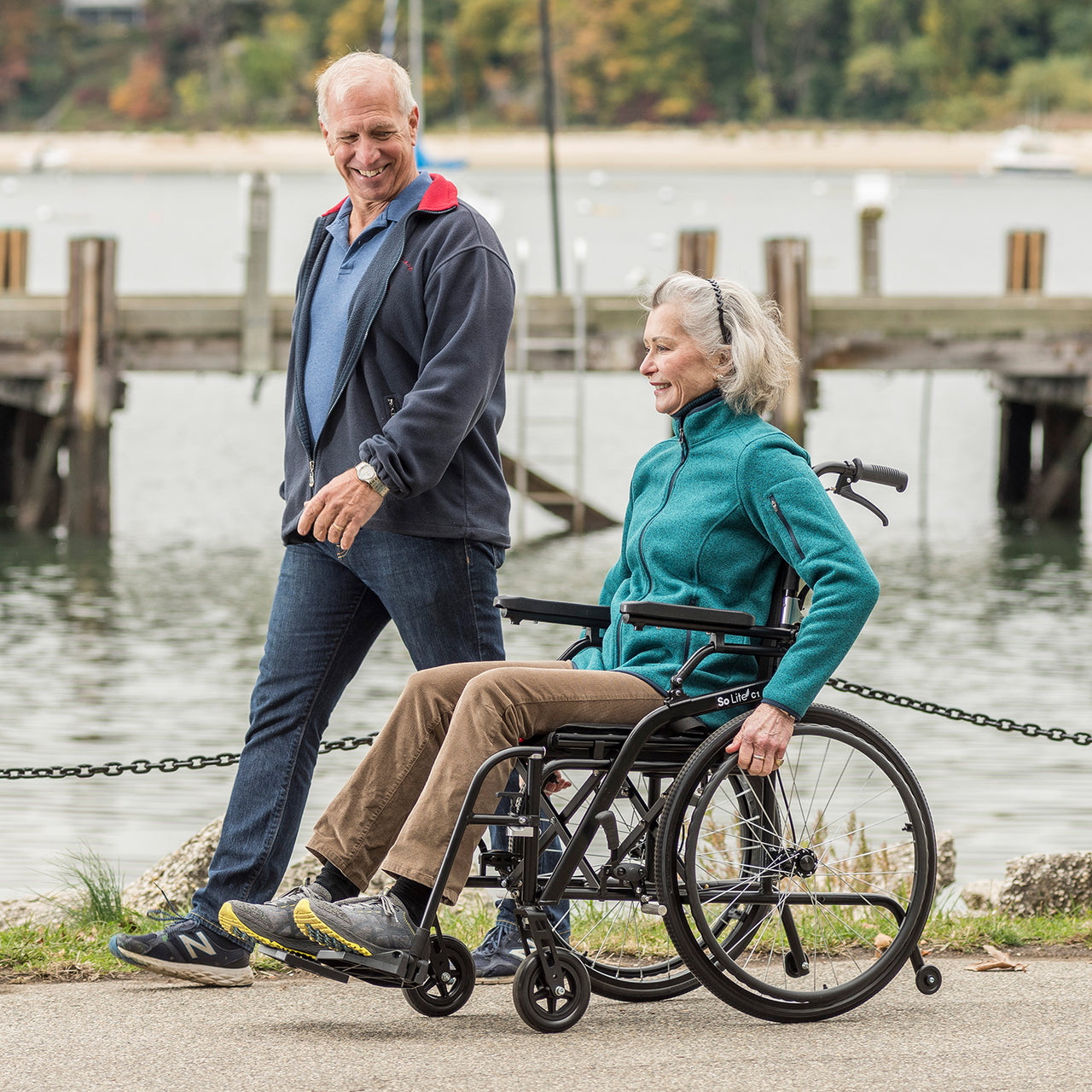 An older woman in a Journey So Lite® C2 Ultra Lightweight Wheelchair smiles as an older man pushes her along a waterfront path. Both look happy and casual, enjoying easy mobility with docks and calm water in the background.
