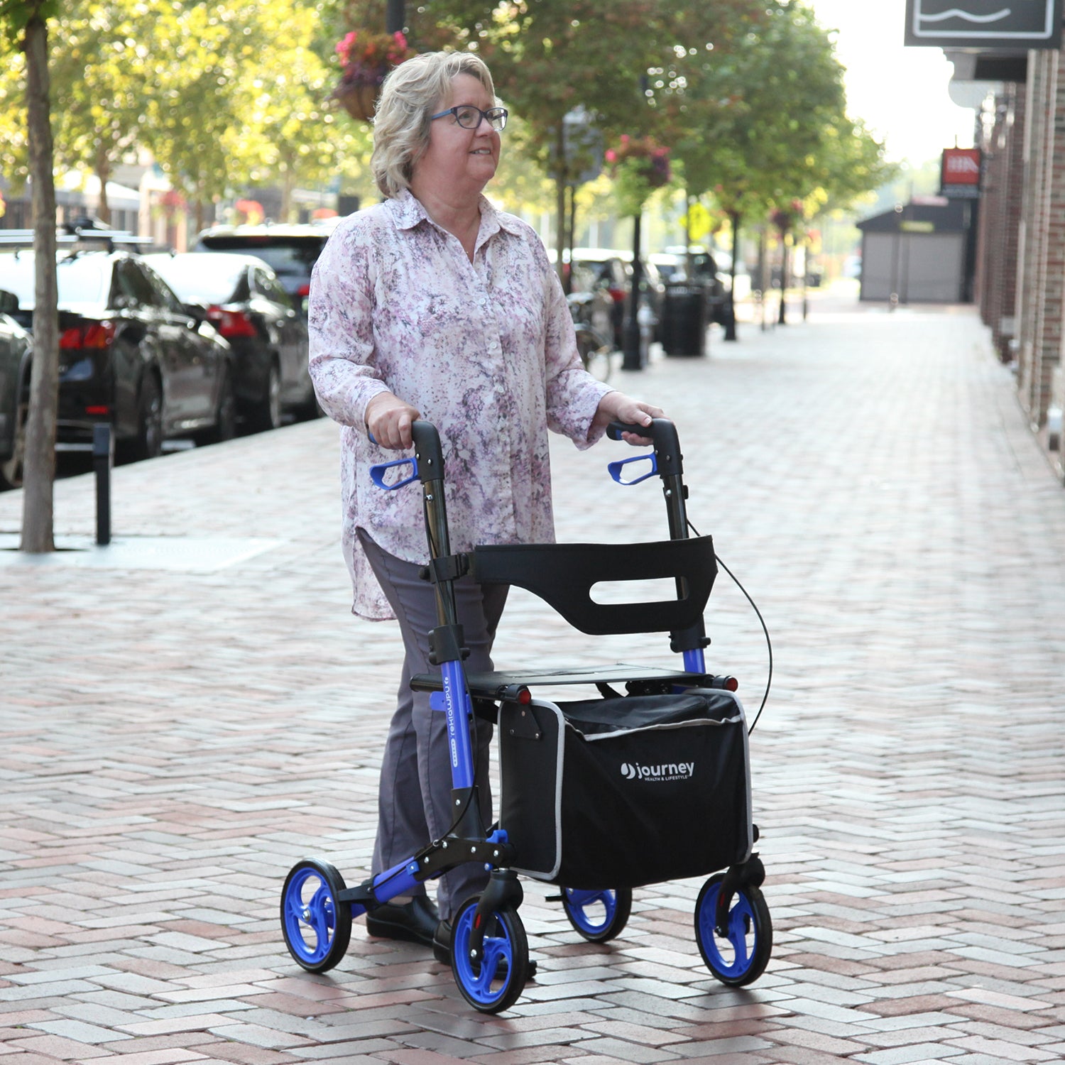 A woman with gray hair, glasses, a floral shirt, and gray pants uses the blue Breeze Rollator with a black storage bag as she walks on a brick sidewalk along a tree-lined city street.