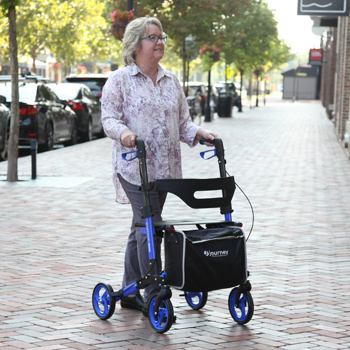 A woman with gray hair, glasses, a floral shirt, and gray pants uses the blue Breeze Rollator with a black storage bag as she walks on a brick sidewalk along a tree-lined city street.