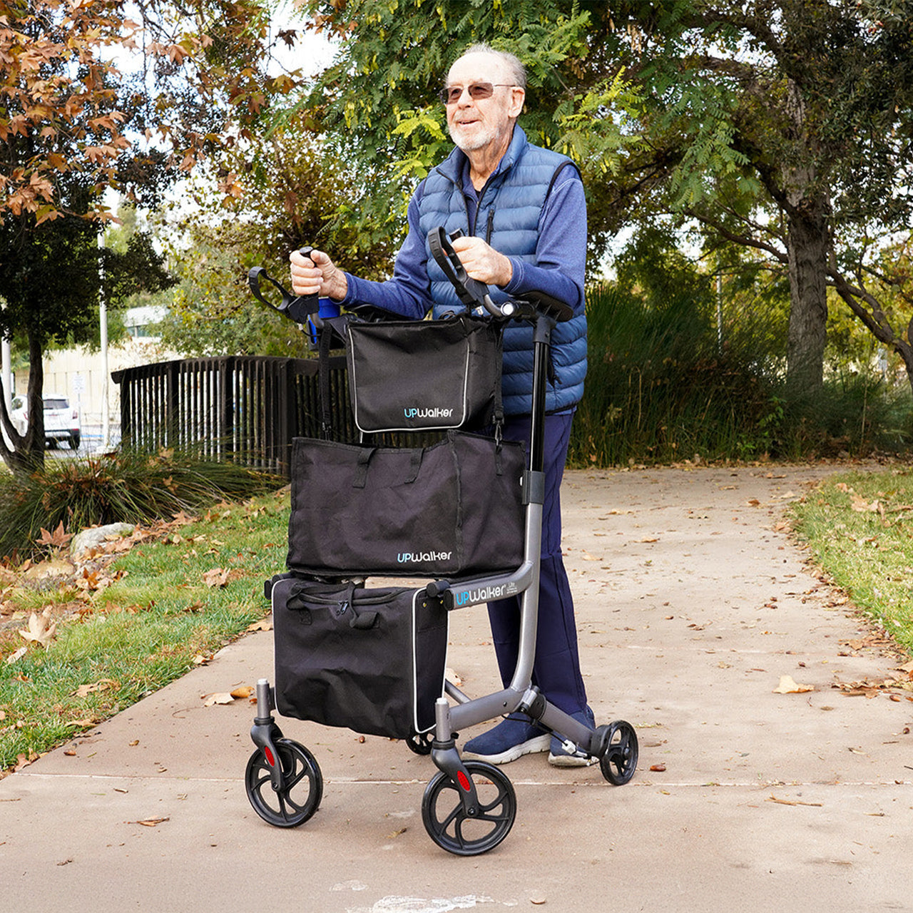 An older man with upright posture, wearing sunglasses and a blue vest, smiles while walking outdoors on a paved path surrounded by grass and trees, using his UPWalker® EZ Lite lightweight mobility aid.