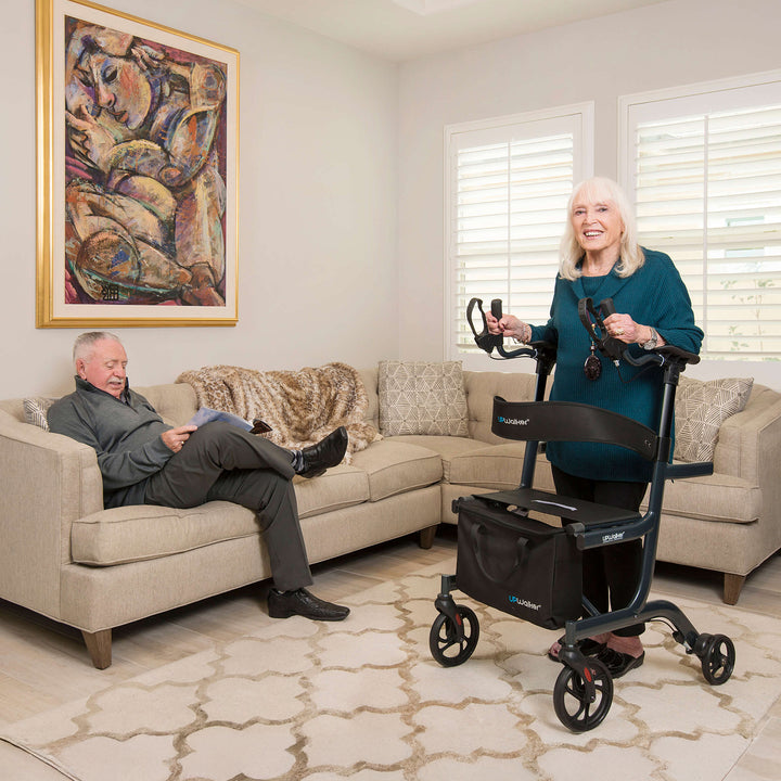 An older woman smiles while standing with her (draft)UPWalker® Lite, a lightweight mobility solution, in a bright living room. An older man reads on a beige sofa as sunlight streams through white shutters onto abstract art.