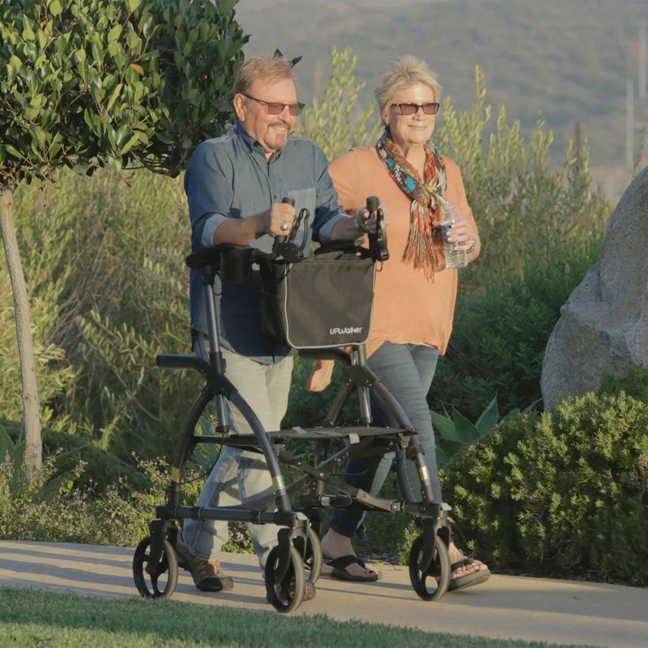 A man uses the UPWalker® Original upright rollator as he walks outdoors with a woman holding a water bottle; they are surrounded by greenery and hills on a sunny day.