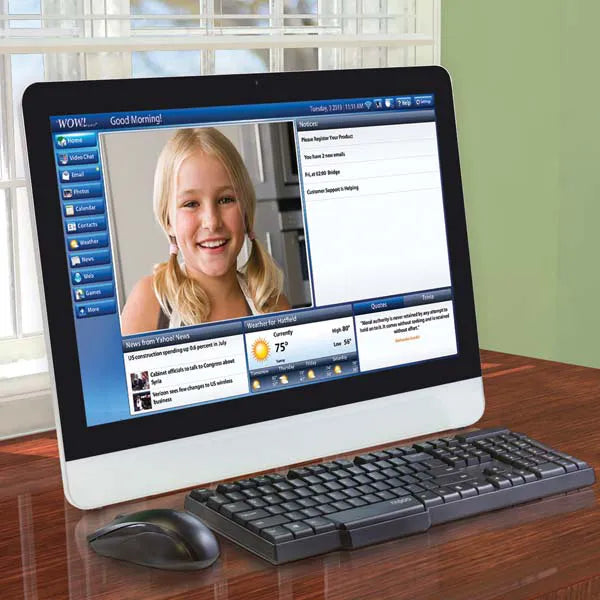 A WOW Computer sits on a wooden desk, displaying a video call with a smiling young girl. The easy-to-use screen also shows news, weather info, and menu options, with a keyboard and mouse in front.