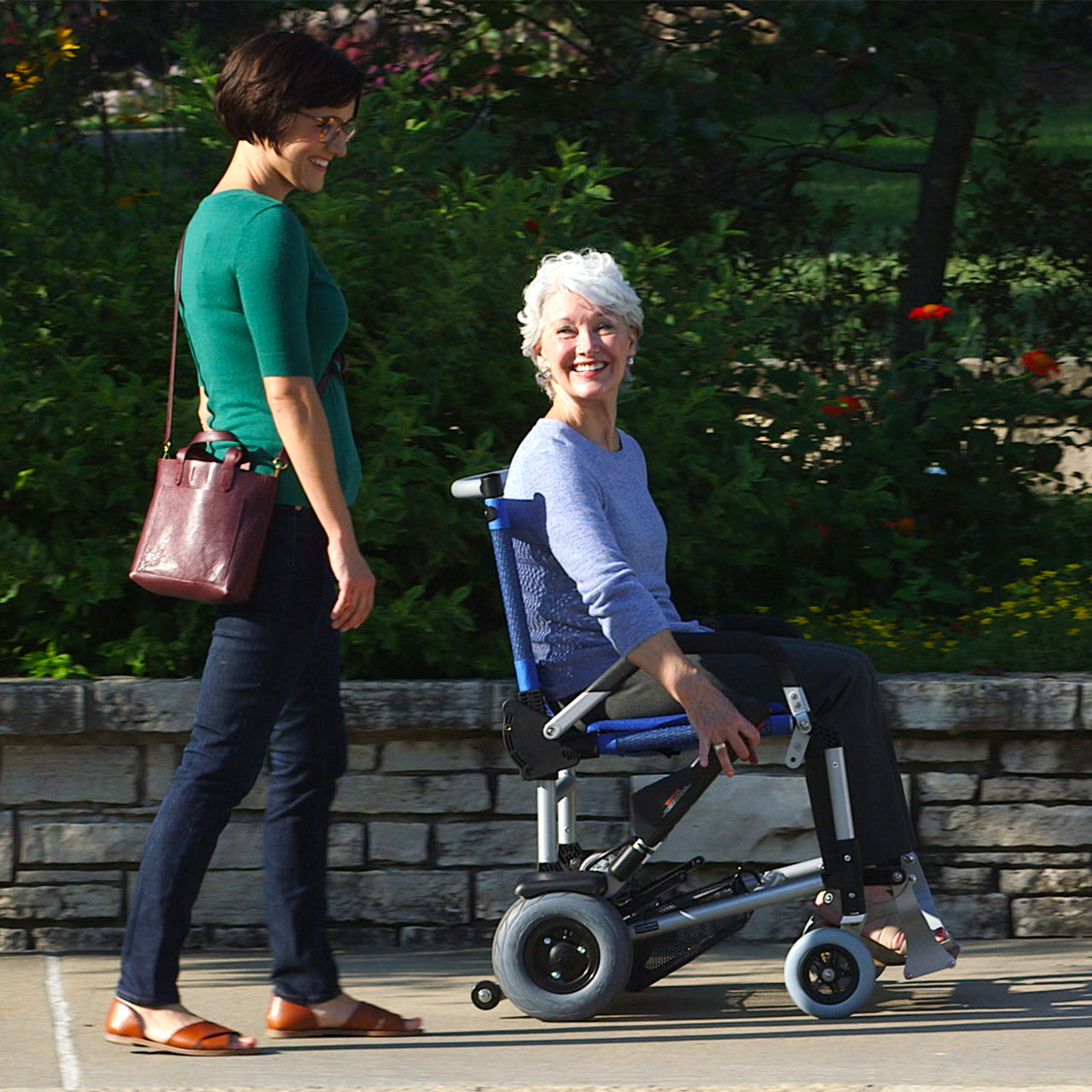 Two women are outdoors on a sunny day; one is pushing the other, who has gray hair and is smiling, in a Journey Zinger® Folding Power Chair along a paved path beside a stone wall and garden with green plants and flowers.