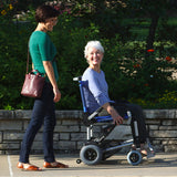 Two women are outside on a sunny day; one smiles while sitting in a Journey Zinger® Folding Power Chair, as the other walks beside her. They pass blooming flowers and a stone wall in the background.