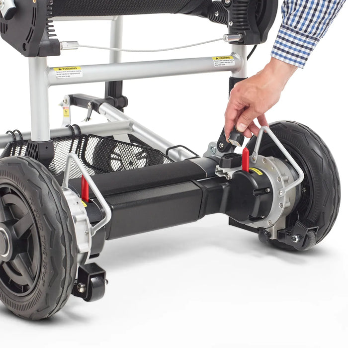 A hand adjusts a red lever near the rear wheel of the Journey Zoomer Lightweight Folding Power Chair, demonstrating its brake release or locking mechanism. The frame and wheels are visible.