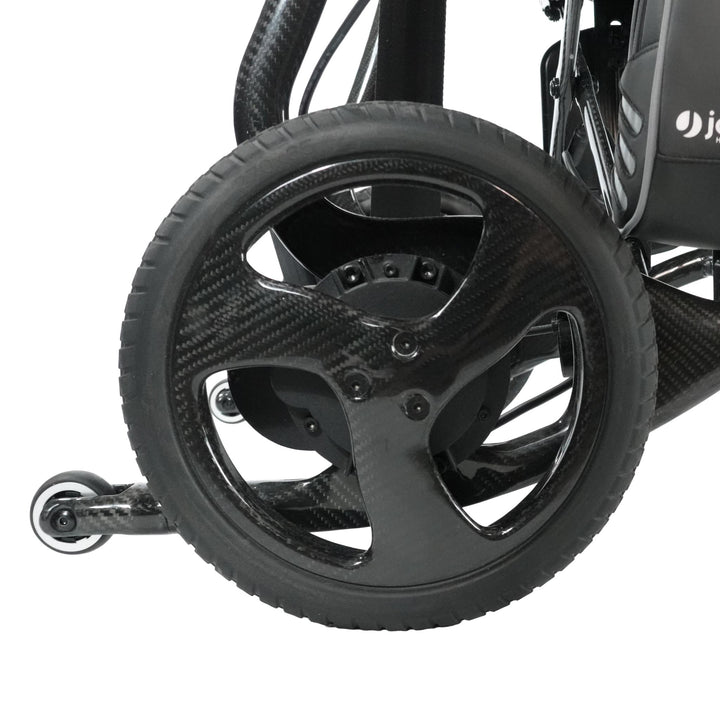 Close-up of the Journey Air Elite Max's black carbon fiber wheel attached to its lightweight frame, with a small caster wheel in the background, highlighting the wheelchair’s sleek, modern design and textured carbon fiber construction.