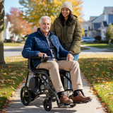 An older man smiles while seated in a Journey Air Elite Max Ultra-Lightweight Portable Power Chair on a sunny sidewalk, as a younger woman in a coat and beanie stands behind him. Autumn leaves and suburban homes can be seen in the background.