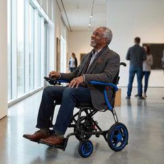 A man smiles as he navigates an art gallery in the Journey Certified Pre-Owned Air Elite Lightweight Power Chair Red. Large windows, people, and colorful artworks create a vibrant, welcoming scene in the background.