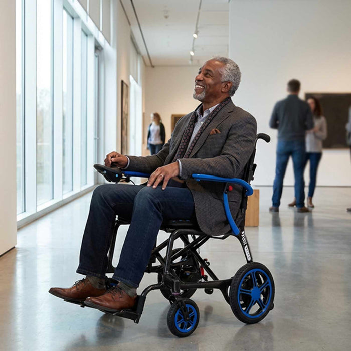 A man smiles as he navigates an art gallery in the Journey Certified Pre-Owned Air Elite Lightweight Power Chair Red. Large windows, people, and colorful artworks create a vibrant, welcoming scene in the background.