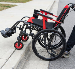 Only the lower legs of a person are visible as they help the Journey So Lite® C2 Ultra Lightweight Wheelchair up a sidewalk curb; its foldable frame has front wheels on the curb while rear wheels remain on the street.