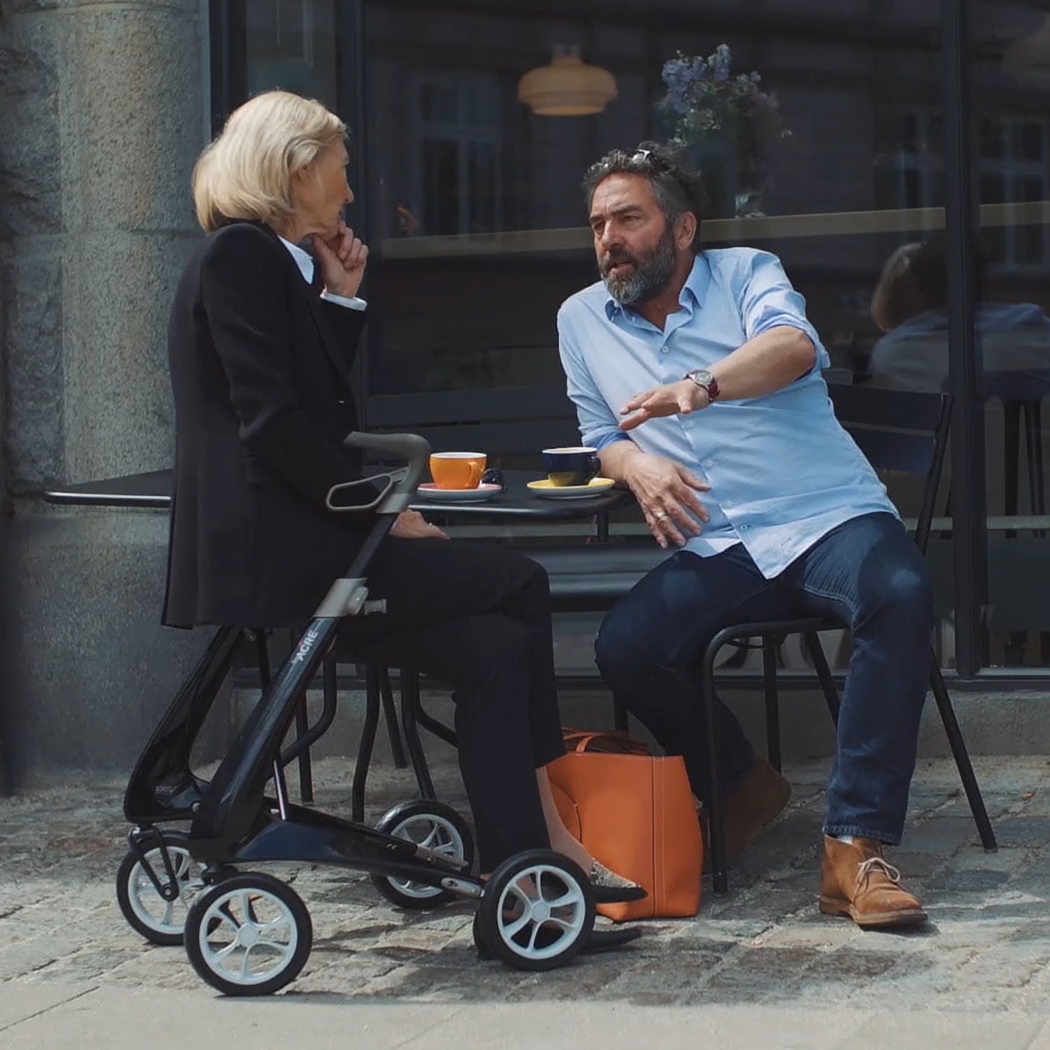A woman sits with a man at an outdoor café table, chatting over coffee. Next to her is the byACRE Carbon Ultralight Rollator and an orange bag, with a building facade visible in the background.