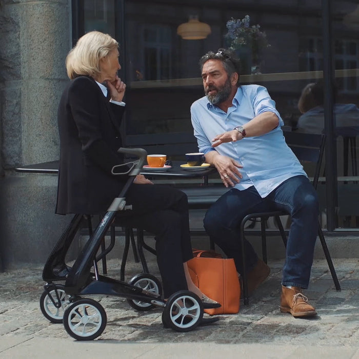 A woman sits with a man at an outdoor café table, chatting over coffee. Next to her is the byACRE Carbon Ultralight Rollator and an orange bag, with a building facade visible in the background.