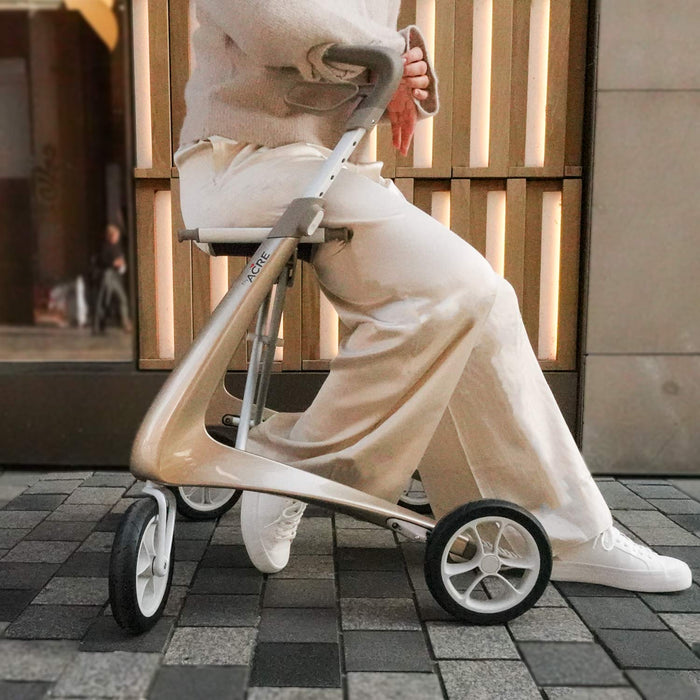 A person in light-colored clothing sits on a byACRE Carbon Ultralight Rollator with a seat, outdoors on a tiled sidewalk near a building.
