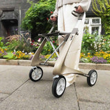 A person in white pants holds a byACRE Carbon Ultralight Rollator with large wheels, standing on a paved path beside a vibrant flower garden and a dark building in the background.