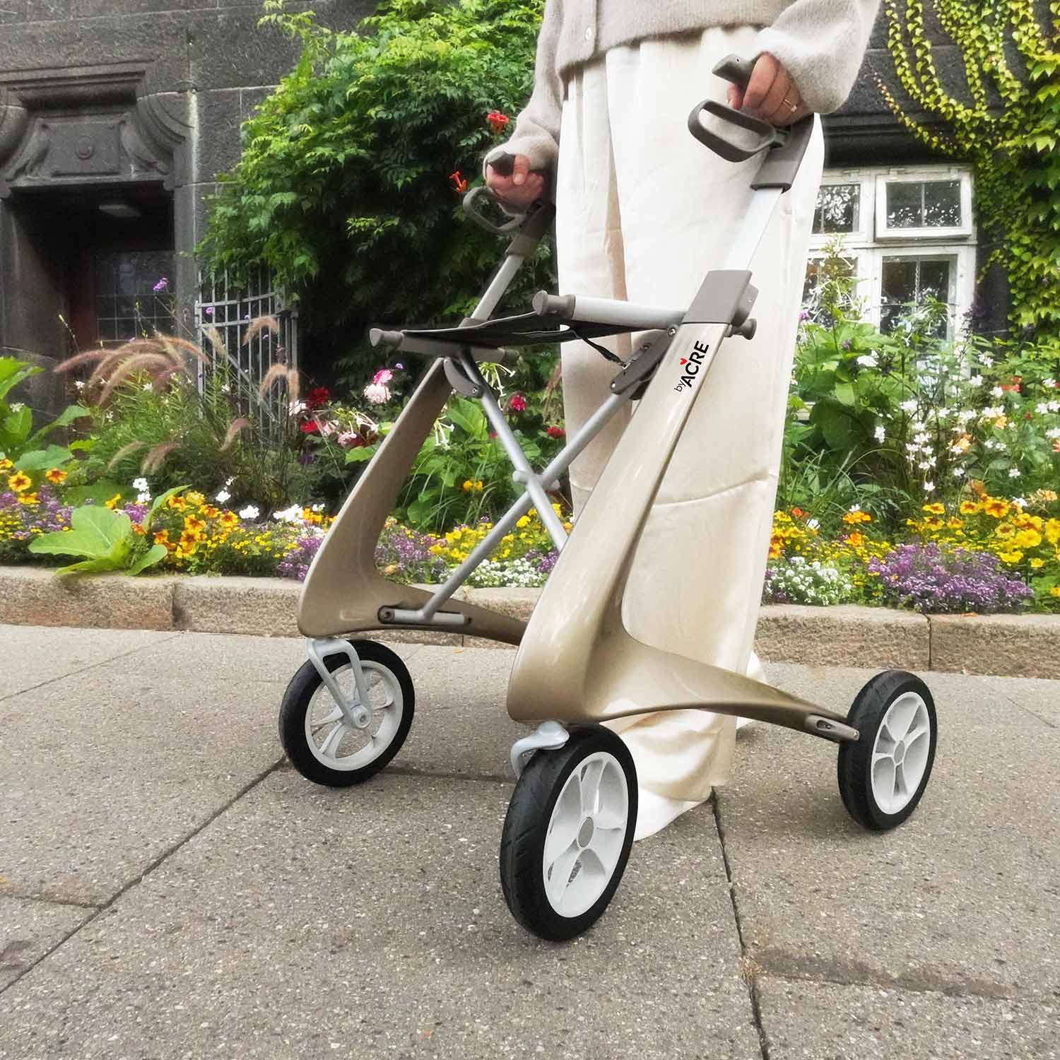 A person in white pants holds a byACRE Carbon Ultralight Rollator with large wheels, standing on a paved path beside a vibrant flower garden and a dark building in the background.