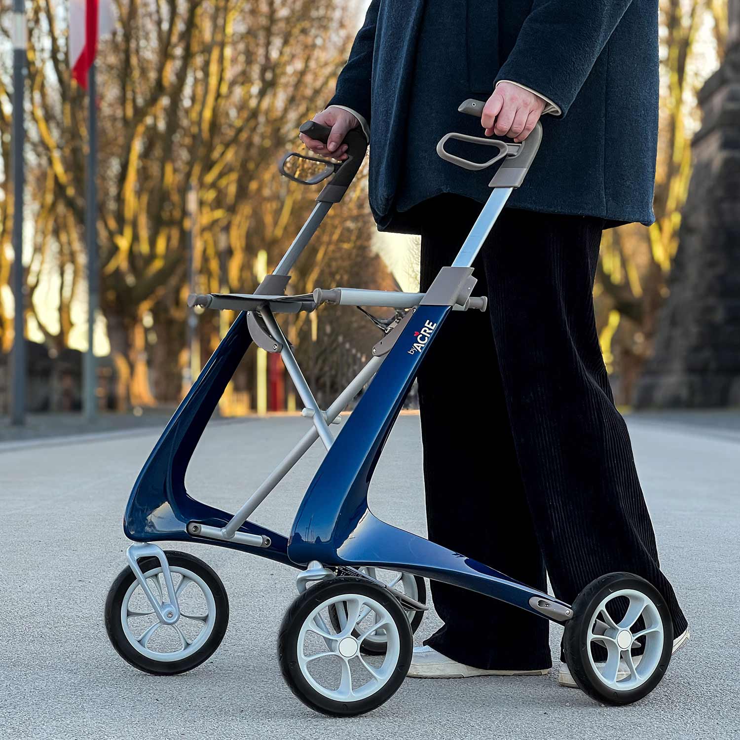 A person in dark clothing uses a blue and silver byACRE Carbon Ultralight Rollator with four wheels on an outdoor path lined with trees.