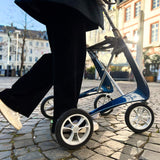 A person in black pants and white shoes walks on a cobblestone street using a byACRE Carbon Ultralight Rollator, with buildings and trees in the background.