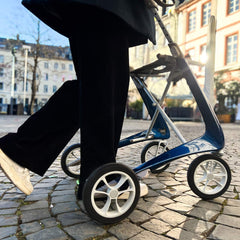 A person in black pants and white shoes walks on a cobblestone street using a byACRE Carbon Ultralight Rollator, with buildings and trees in the background.